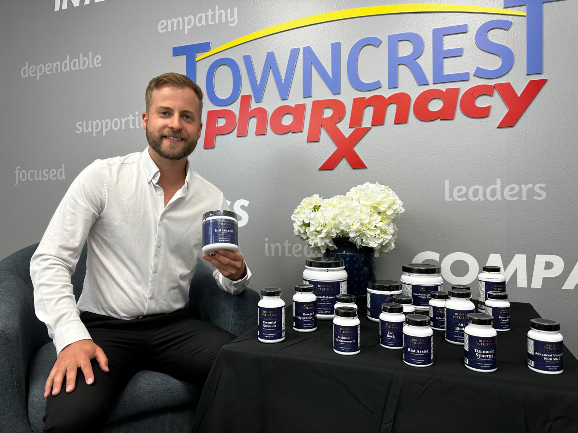 Aaron, the Towncrest pharmacist, seated in an armchair and holding a supplement tub, with a table of Rooted Intuition products beside him